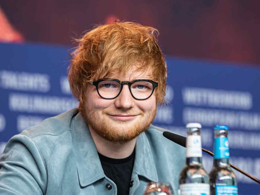 A portrait of Ed Sheeran smiling benignly, with a mop of professionally tousled red har and black-framed glasses.